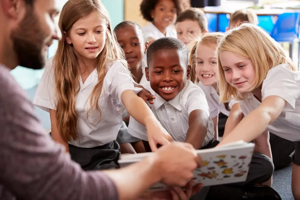 teacher is sitting surrounded by children reading a book